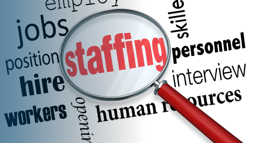 A graphic illustration of a magnifying glass with a red handle zooming in on the word "staffing" in bold red text. Surrounding the magnifying glass on a light blue and white gradient background are related workforce terms in dark blue and black text, including "jobs," "position," "hire," "workers," "employ," "open," "skills," "personnel," "interview," and "human resources."