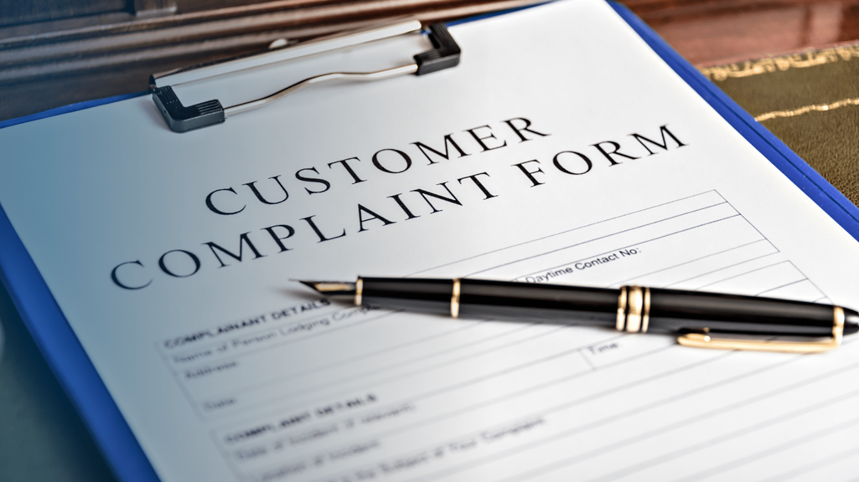 A close-up of a "Customer Complaint Form" on a blue clipboard, with fields for complainant details, complaint details, and a daytime contact number. A black and gold fountain pen rests diagonally across the form. The clipboard is placed on a wooden surface.