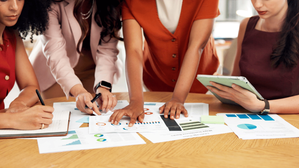 Four women lean over a wooden conference table covered in printed data reports featuring bar charts, pie charts, and line graphs. One woman writes notes, another points to a chart with a pen, a third leans in to review the documents, and a fourth holds a tablet. The women are dressed in professional attire in shades of red, pink, orange, and burgundy. Only their torsos and hands are visible.
