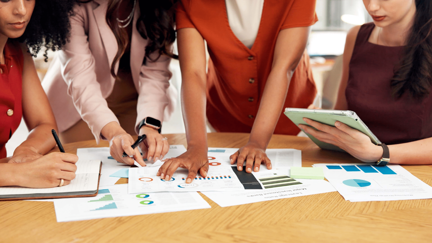 Four women lean over a wooden conference table covered in printed data reports featuring bar charts, pie charts, and line graphs. One woman writes notes, another points to a chart with a pen, a third leans in to review the documents, and a fourth holds a tablet. The women are dressed in professional attire in shades of red, pink, orange, and burgundy. Only their torsos and hands are visible.