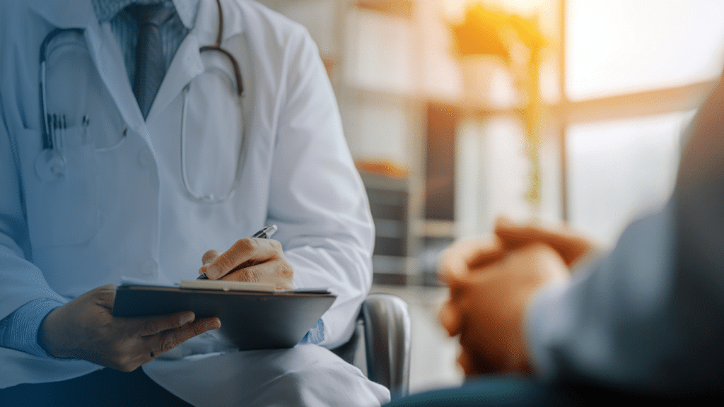 A close-up of a doctor in a white lab coat and stethoscope writing notes on a clipboard during a patient consultation. The patient, shown blurred in the foreground with hands clasped, sits across from the doctor. A warmly lit room with a plant is softly visible in the background. Only the torsos and hands of both individuals are visible.