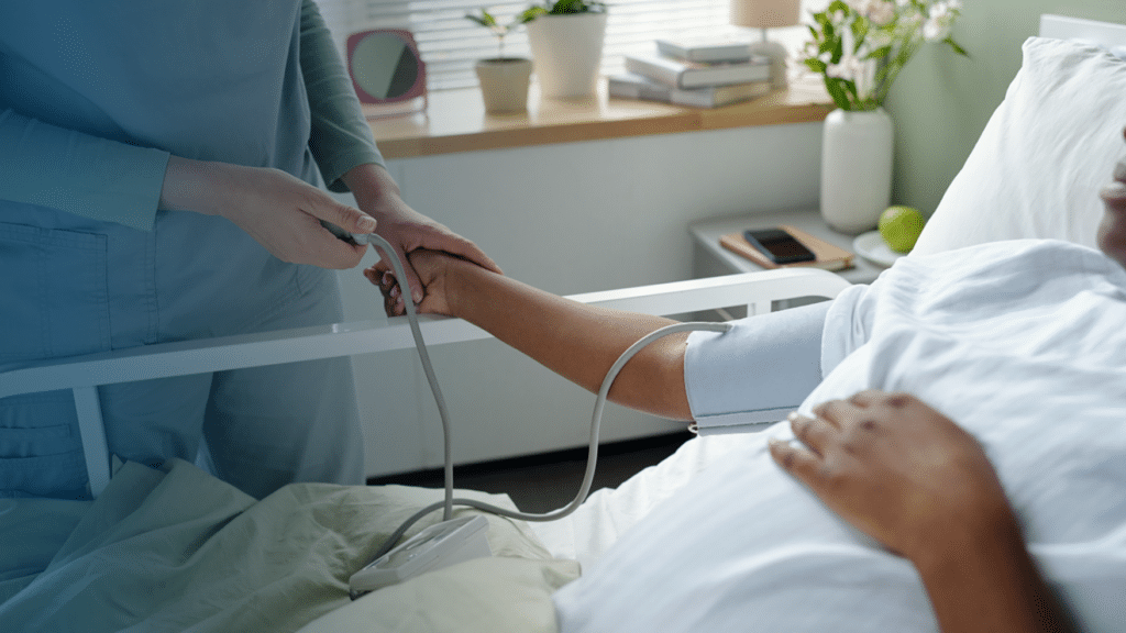 A healthcare worker in teal scrubs places a pulse oximeter on the finger of a patient lying in a hospital bed with a blood pressure cuff on their arm. The patient is dressed in a white hospital gown and resting under white sheets. A bedside table in the background holds a potted plant, stacked books, a phone, a green apple, and a vase of pink flowers. Only the torsos and hands of both individuals are visible.