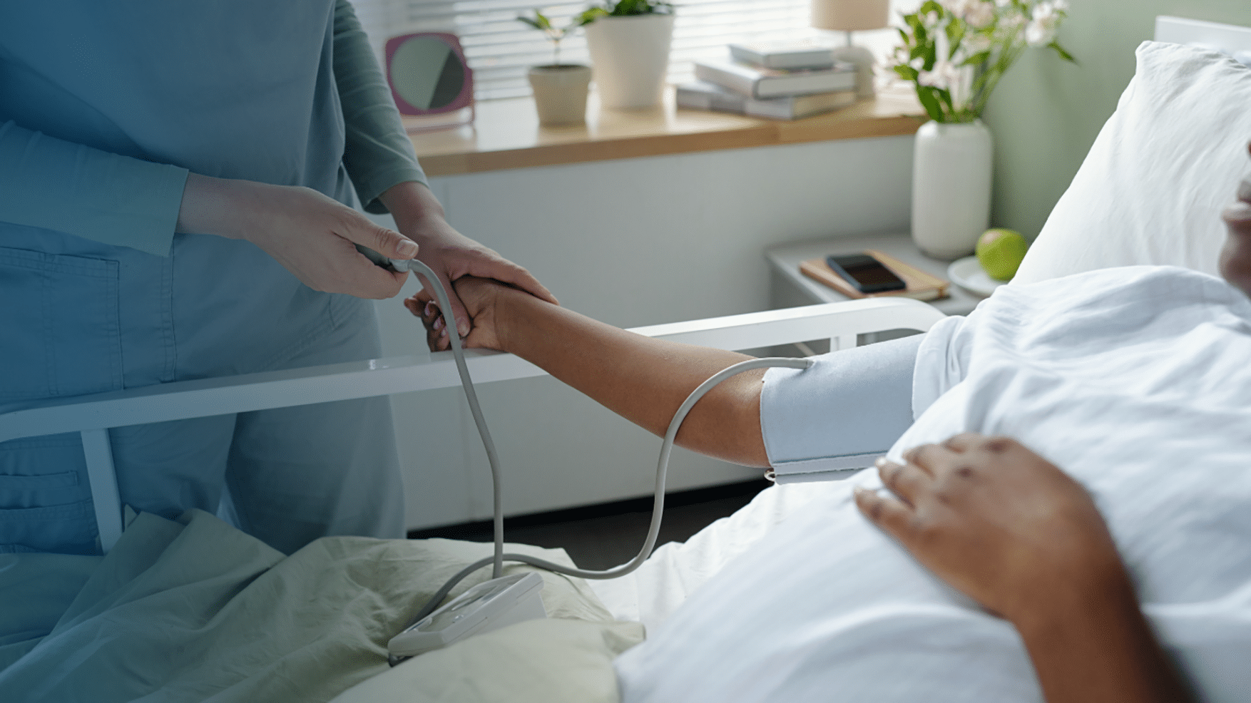 A healthcare worker in teal scrubs places a pulse oximeter on the finger of a patient lying in a hospital bed with a blood pressure cuff on their arm. The patient is dressed in a white hospital gown and resting under white sheets. A bedside table in the background holds a potted plant, stacked books, a phone, a green apple, and a vase of pink flowers. Only the torsos and hands of both individuals are visible.