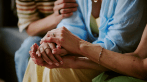 A close-up of three people seated together, with two sets of hands clasped together in a gesture of comfort and support. One person wearing a light blue shirt and a delicate chain bracelet gently holds the hands of another person wearing rings and a yellow skirt. In the background, a third person in a striped shirt rests their hands on the shoulder of the person in blue. Only the torsos and hands of the individuals are visible.